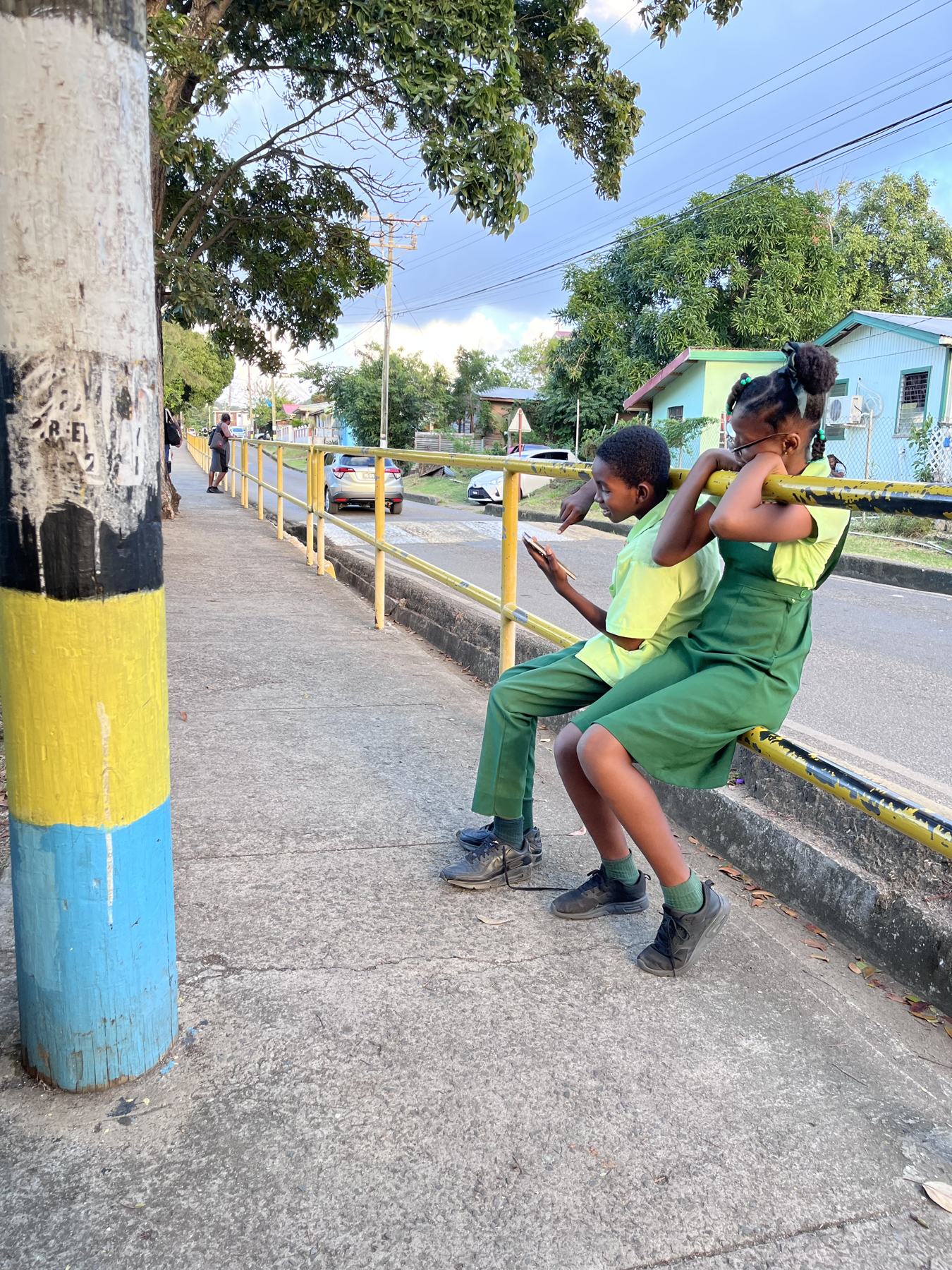 Schoolchildren in green uniforms sitting on a roadside rail next to a pole painted in the colours of the Saint Lucian flag